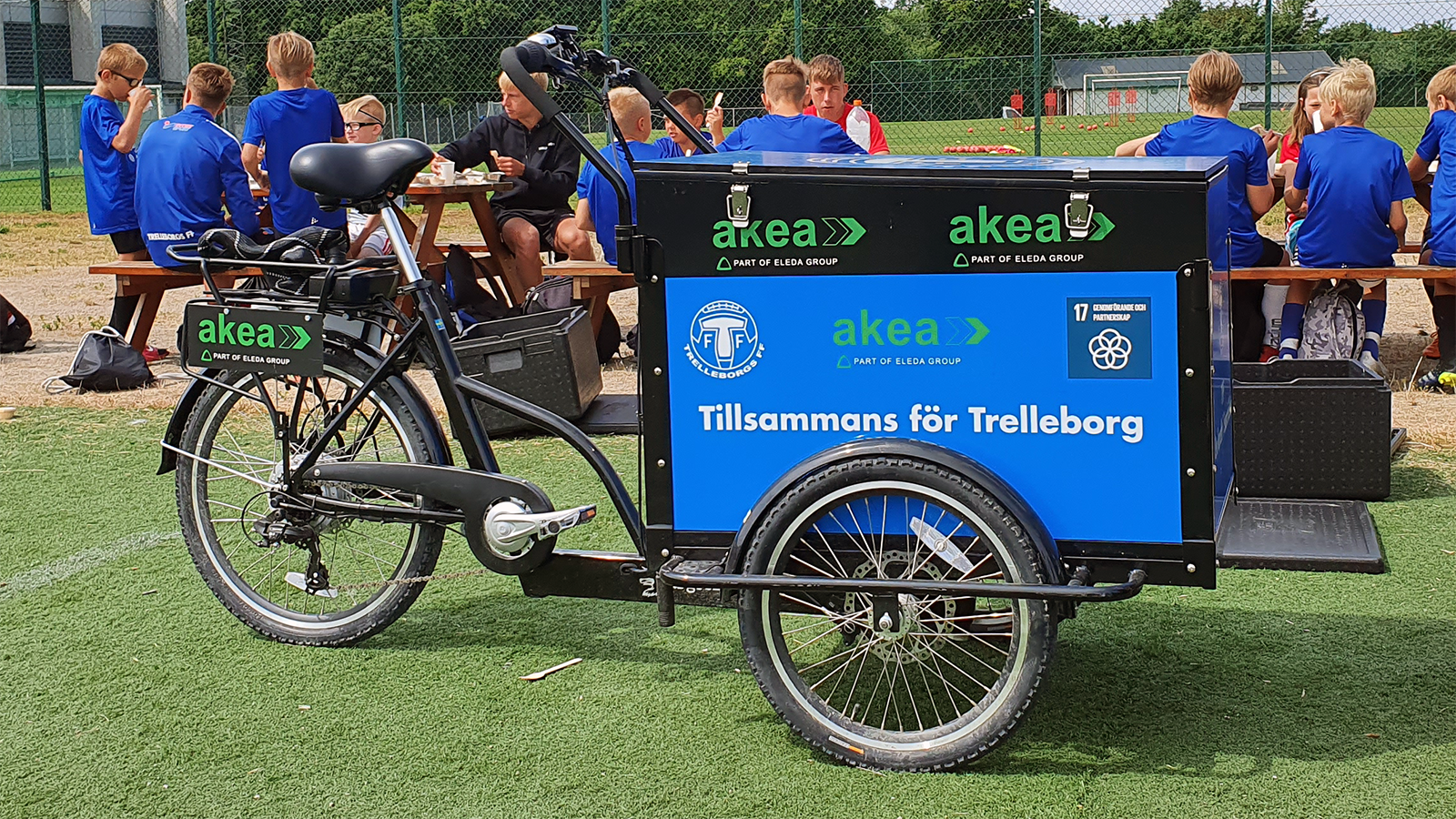 Ungdomsspelare i Trelleborgs FF sitter och äter mellanmål vid träbänkar. I bakgrunden syns en fotbollsplan. I förgrunden, framför spelarna, står en svart lådcykel märkt med Akea och texten "Tillsammans för Trelleborg".